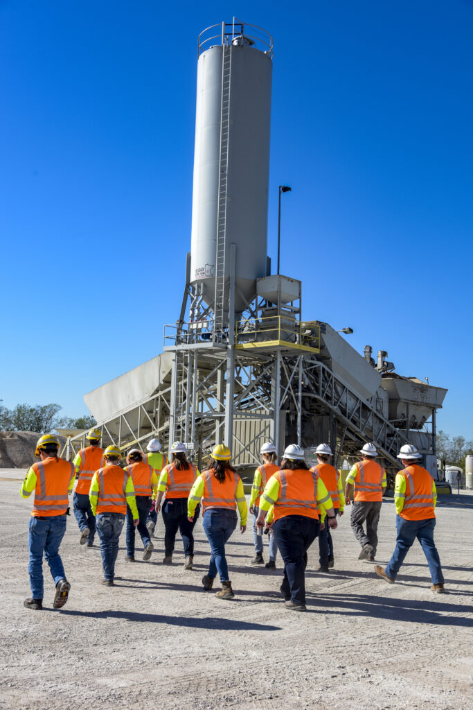 A group of students wearing safety vests and hard hats approach a cement plant.