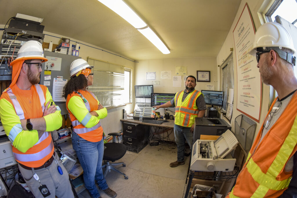 Three individuals in bright vests listen to instructions from a person standing by different types of equipment.