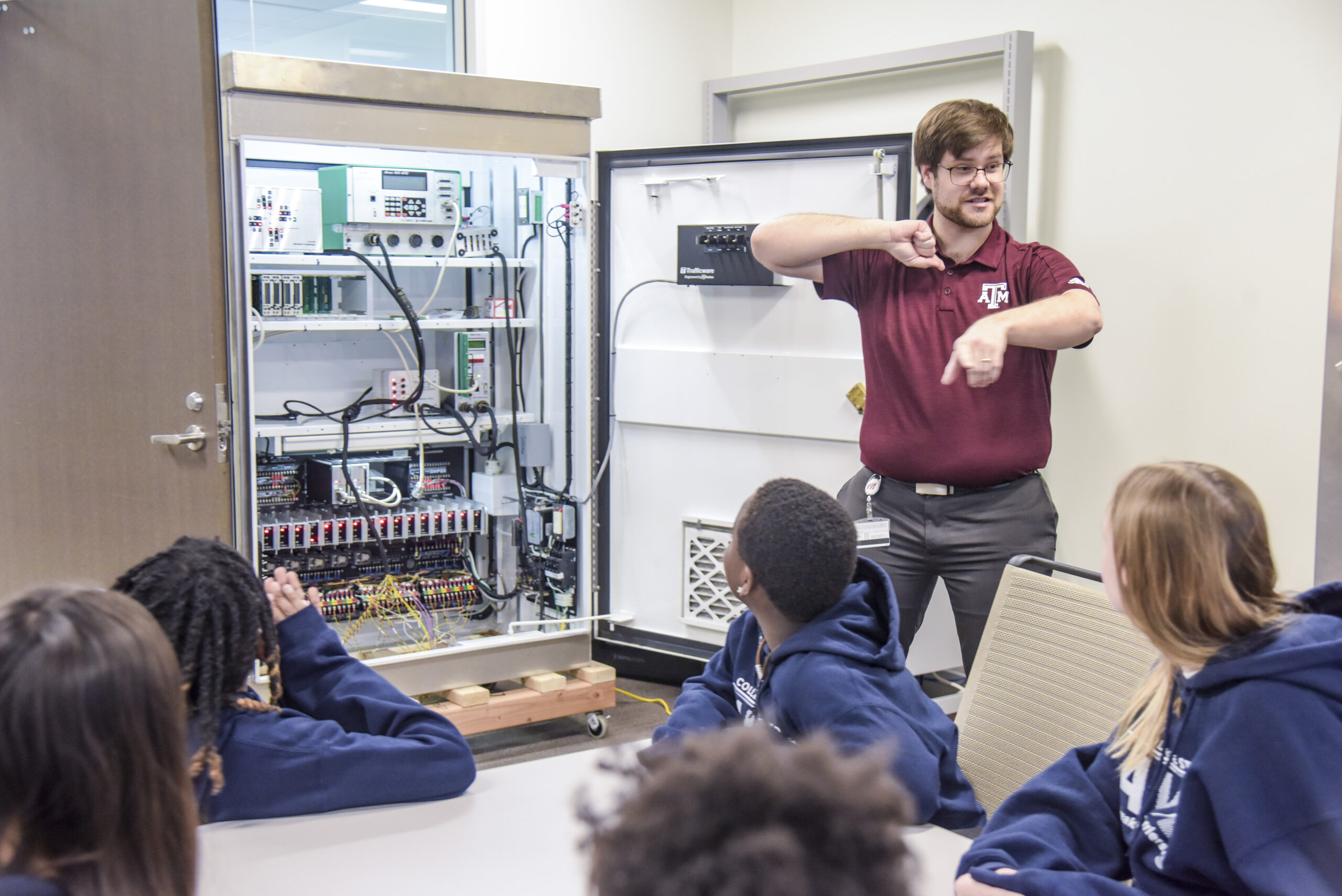 : Instructor stands in front of a group of children explaining the components of a machine.